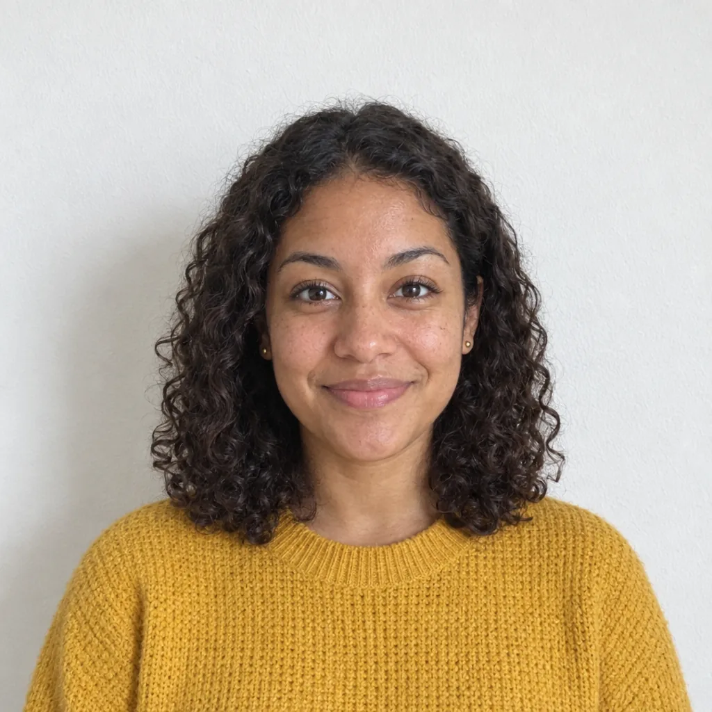 A smiling, curly-haired woman in a mustard knit sweater, photographed against a plain white wall in daylight.