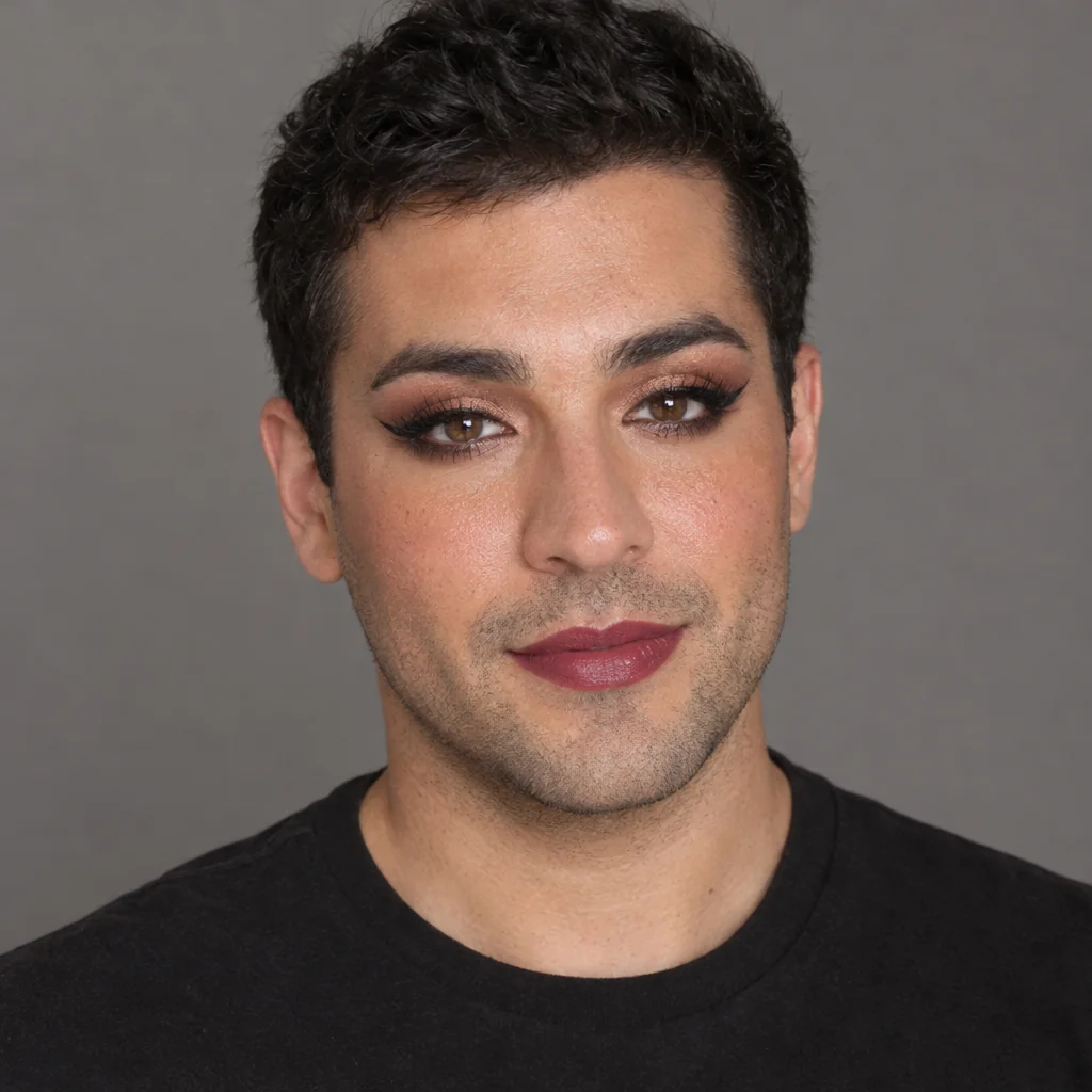 A man with short dark hair, rose-toned lipstick, defined eyes and a black crew-neck top against a grey studio background.
