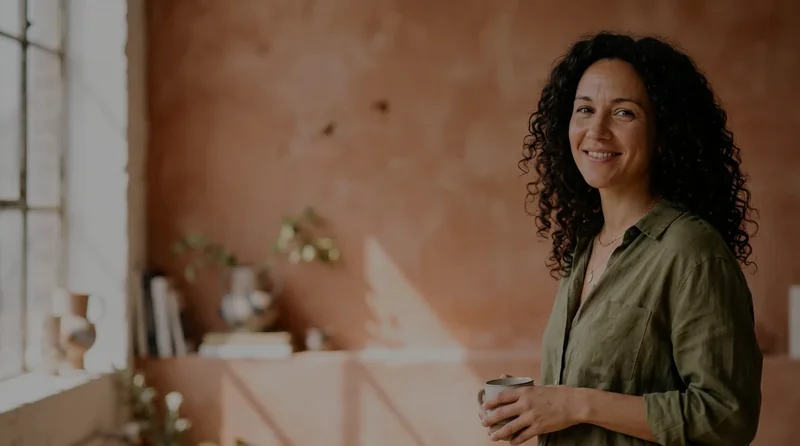 Ein unterbelichtetes Porträt einer Frau mit lockigem Haar und olivgrünem Hemd, die mit einer Tasse am Fenster steht.